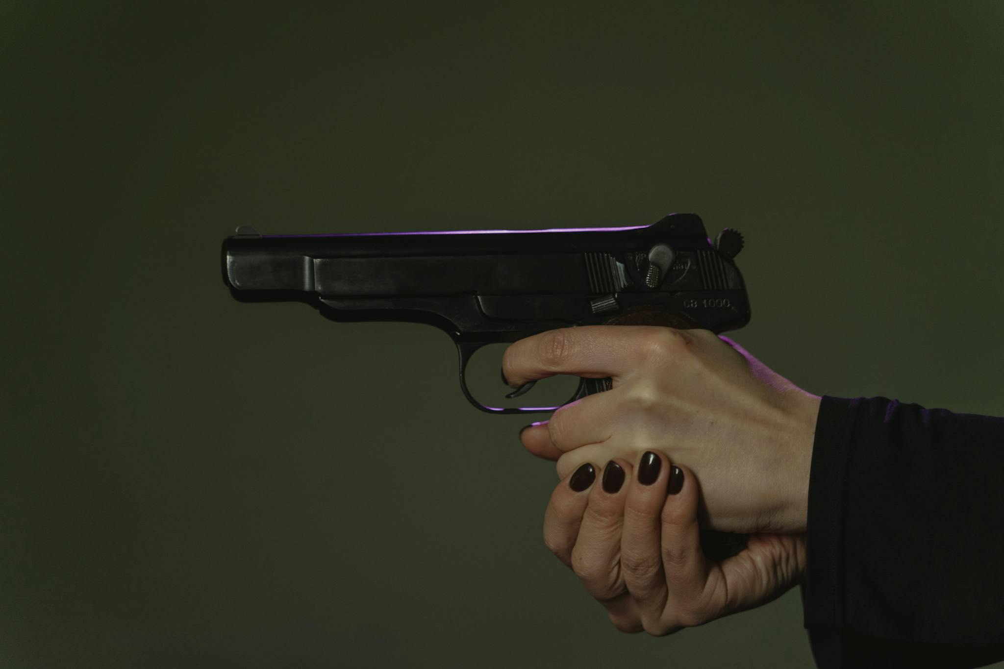 A detailed shot of hands holding a black handgun with dark painted nails.