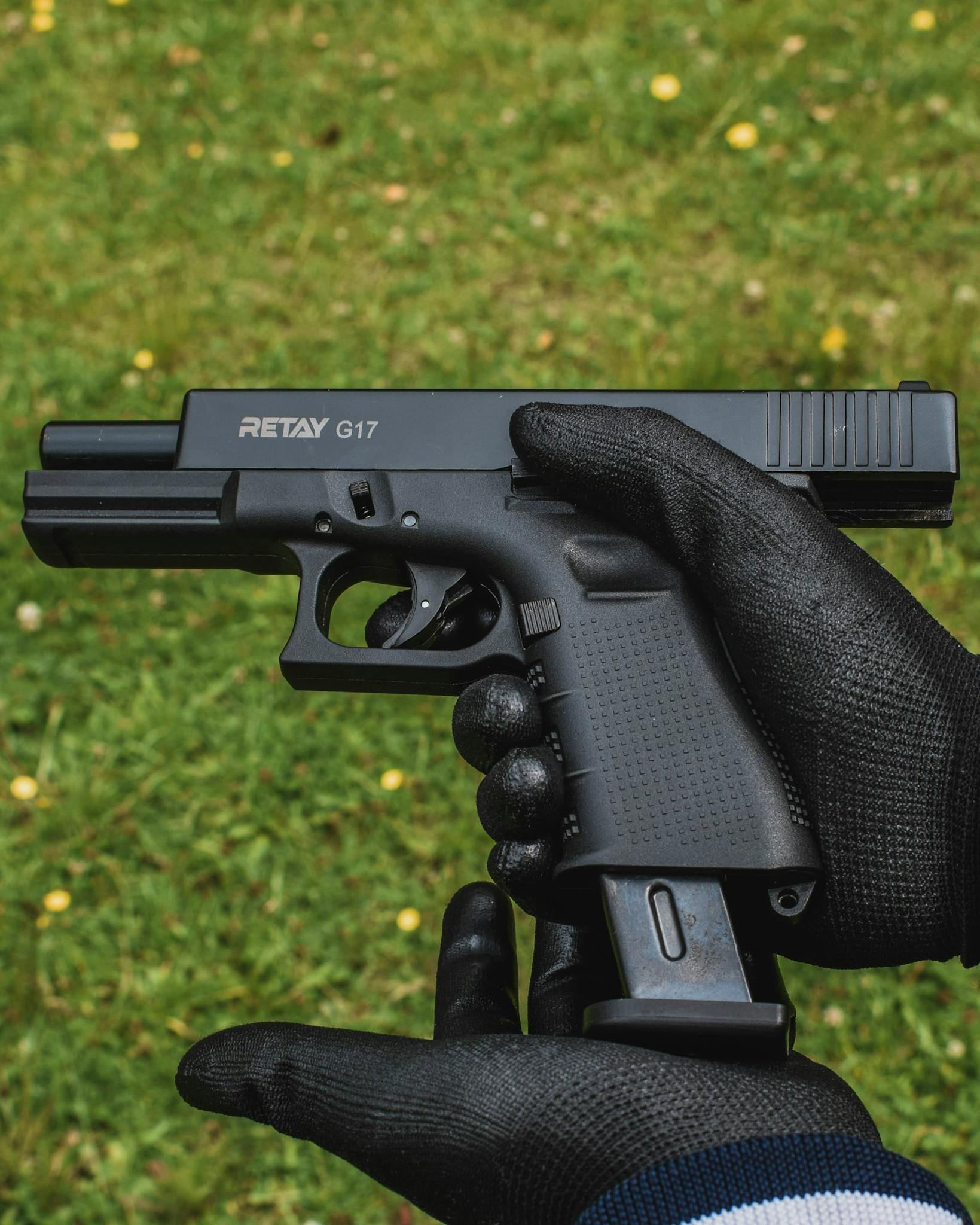 Close-up of a gloved person holding a firearm outdoors on grass.