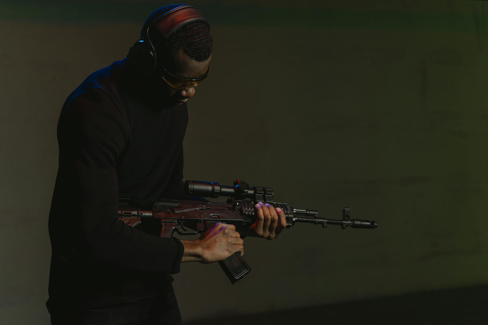Man in black attire focusing intensely while holding a rifle indoors, wearing safety gear.